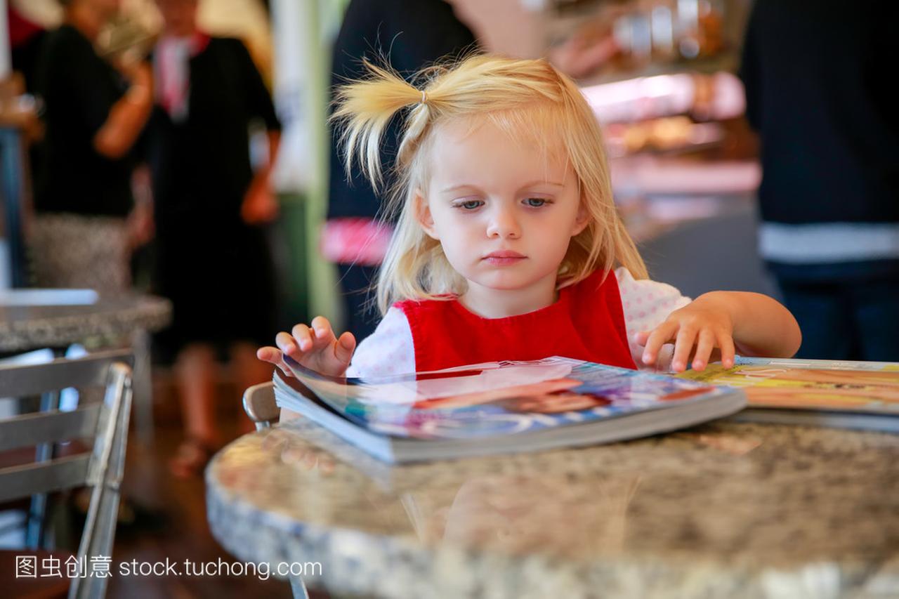 可爱的金发小姑娘看杂志的一家咖啡馆