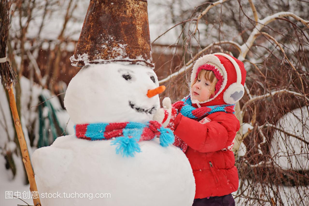 儿童女孩穿着红色的外套,冬天花园里堆雪人