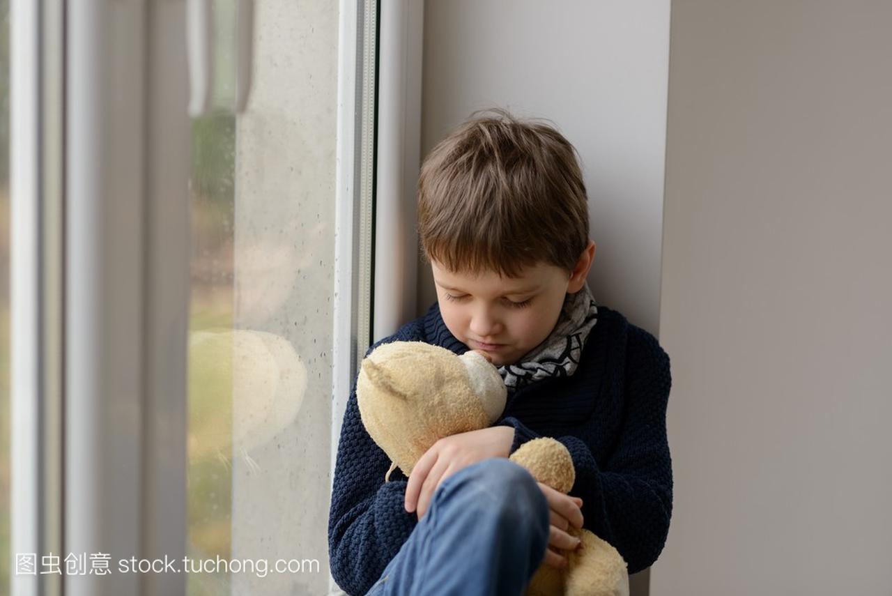 小男孩坐在窗台上和他最喜欢的玩具熊的拥抱