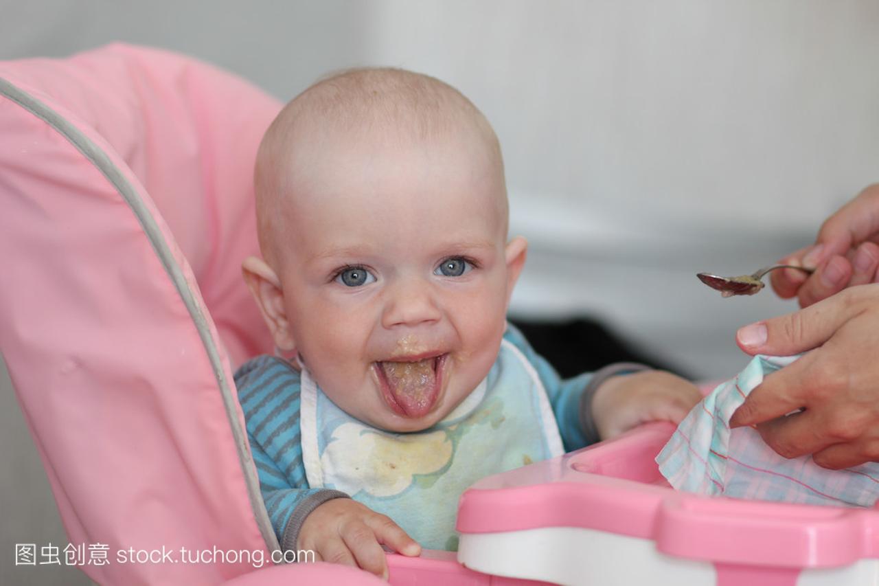 漂亮的宝宝吃粥从妈妈的手。他坐在粉红色的儿