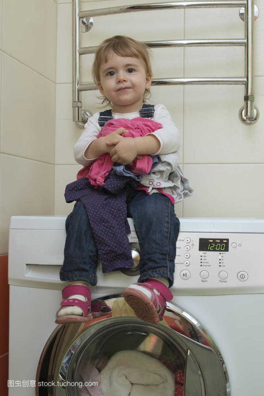 快乐可爱的小女孩坐在洗衣机在家庭内部的衣服
