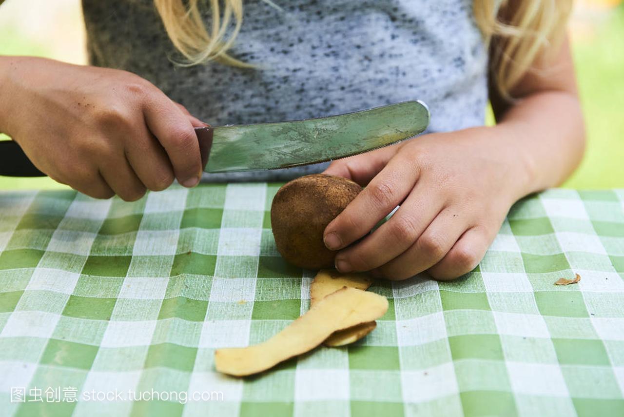 详细的孩子用菜刀手脱皮鲜黄色的土豆。外面的