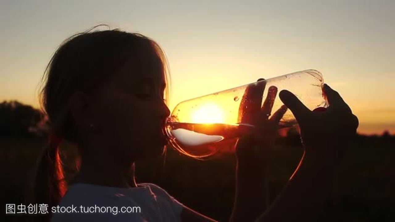 年轻女孩喝水从瓶麦田在夏季日落时的剪影。你