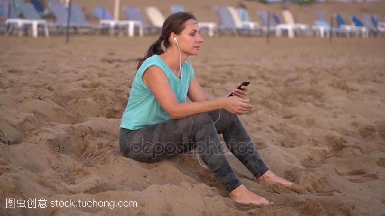 运动妇女与耳机坐在海滩和休息后慢跑。视频在