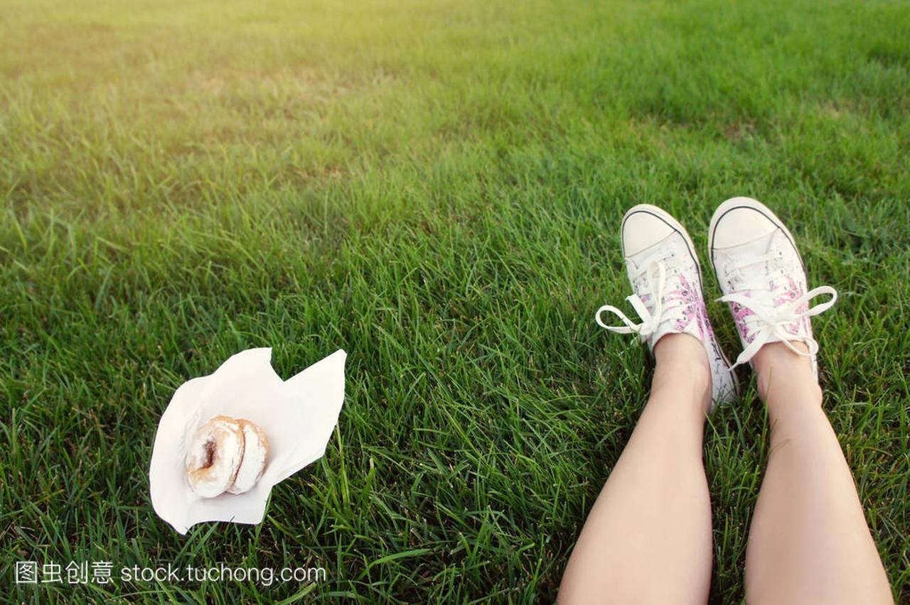 美丽的女性腿在运动鞋和甜面包圈在公园的草