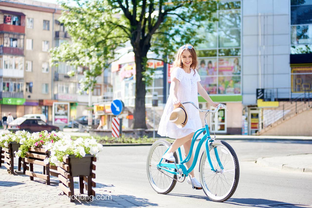 微笑的年轻女性穿着白色礼服骑蓝色自行车在现