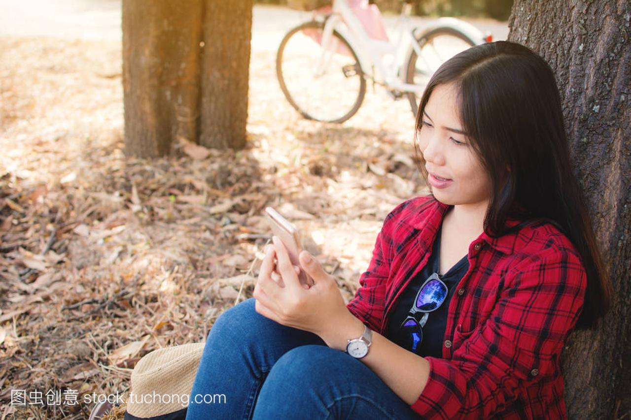 享受片刻的女人使用智能手机坐在公园的大树下