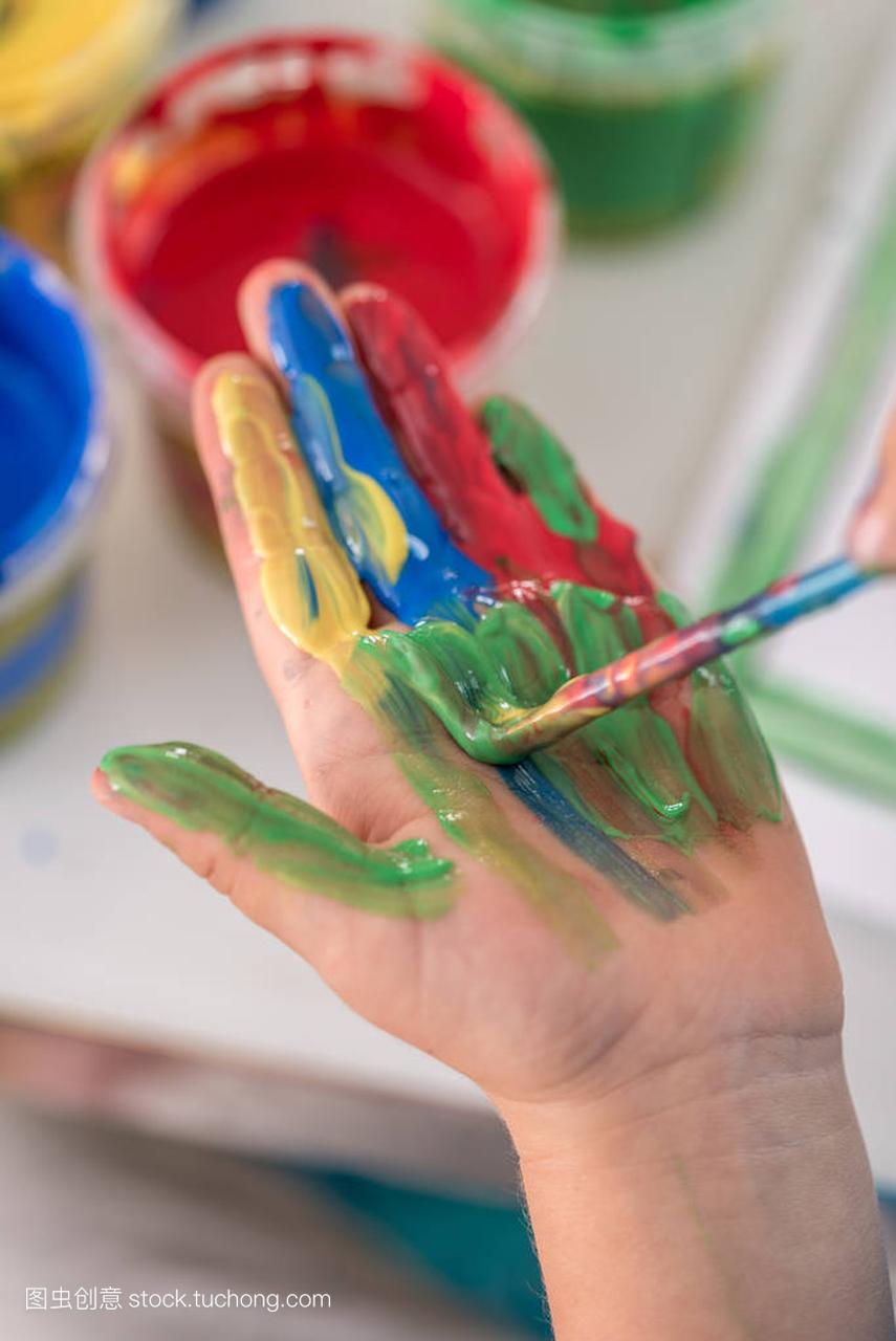 Creative little boy painting its hand with a paintbrush with colorful paint in a close up view of his palm.