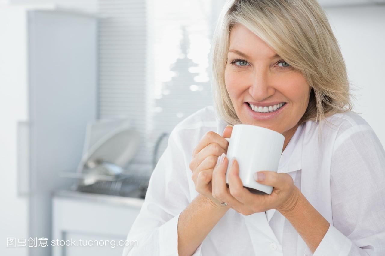 快乐的女人早上喝咖啡,在厨房里看着照相机