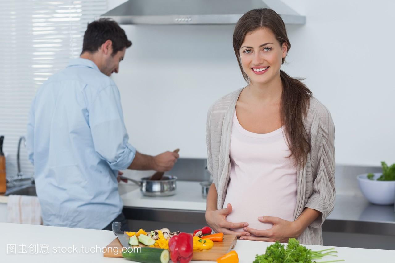 怀孕的女人抱着她的肚子在厨房和她的丈夫在后