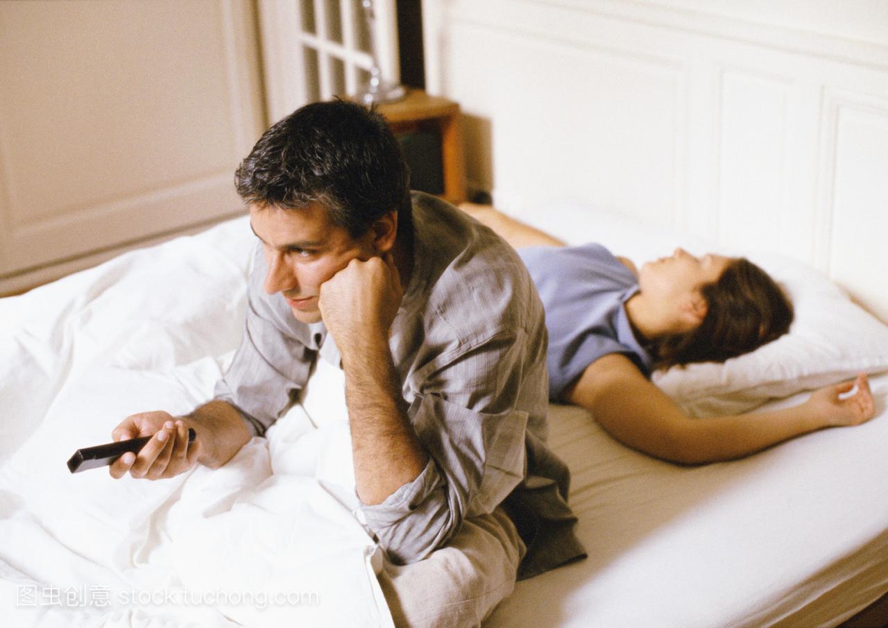 男人坐在床上,指着遥控器,女人躺在他身后。
